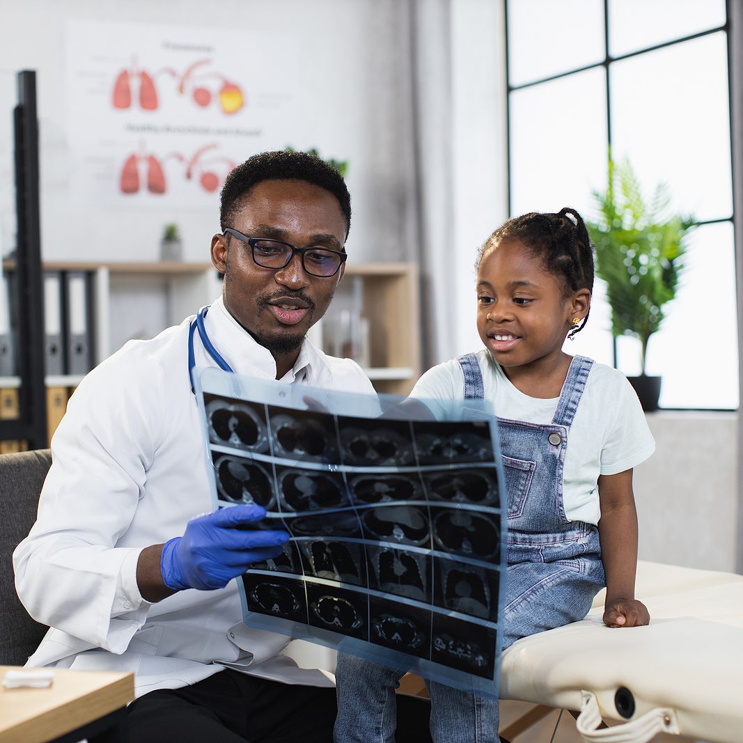 Handsome friendly smiling black-skinned male doctor pediatrician, showing results of x-ray CT scan for little african kid girl in modern medical center during appointment